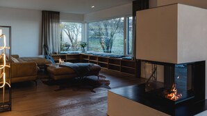 Sala de estar moderna com lareira, piso de madeira, sofá e um assento junto à janela com vista para um jardim.