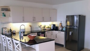 Una cocina moderna con armarios blancos, una encimera negra y un refrigerador de acero inoxidable. Hay sillas blancas alrededor de la isla y una pequeña mesa redonda con estampado floral.