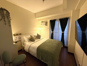 Een hotelkamer met een bed, een stoel, een televisie, een lamp en een raam met gordijnen.