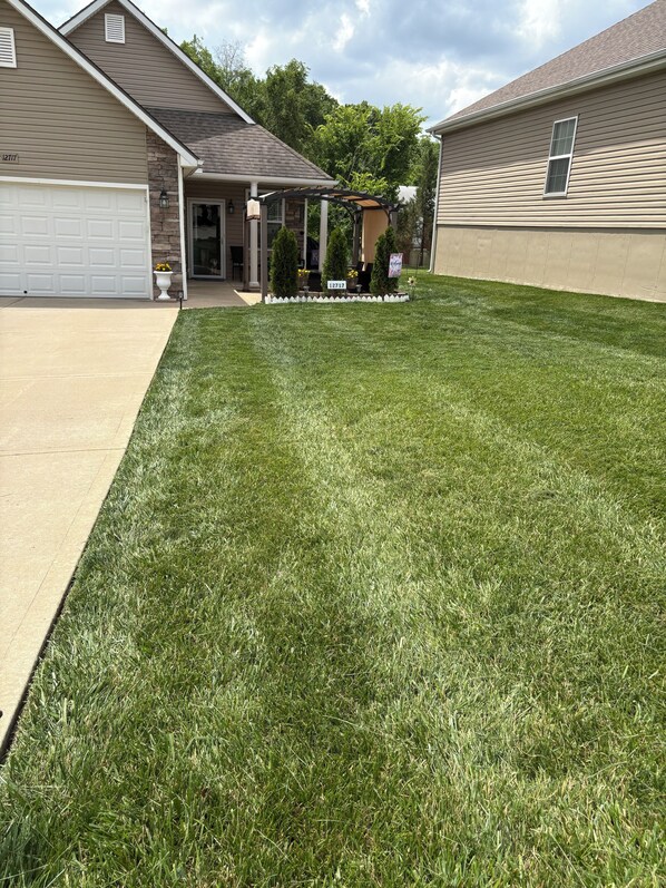 A residential lawn with a garage, a covered patio, and a small garden area with potted plants. - 2 bdrm - Condo in Independence, MO close to Chiefs stadium / World Cup, coolAC, (Independence)