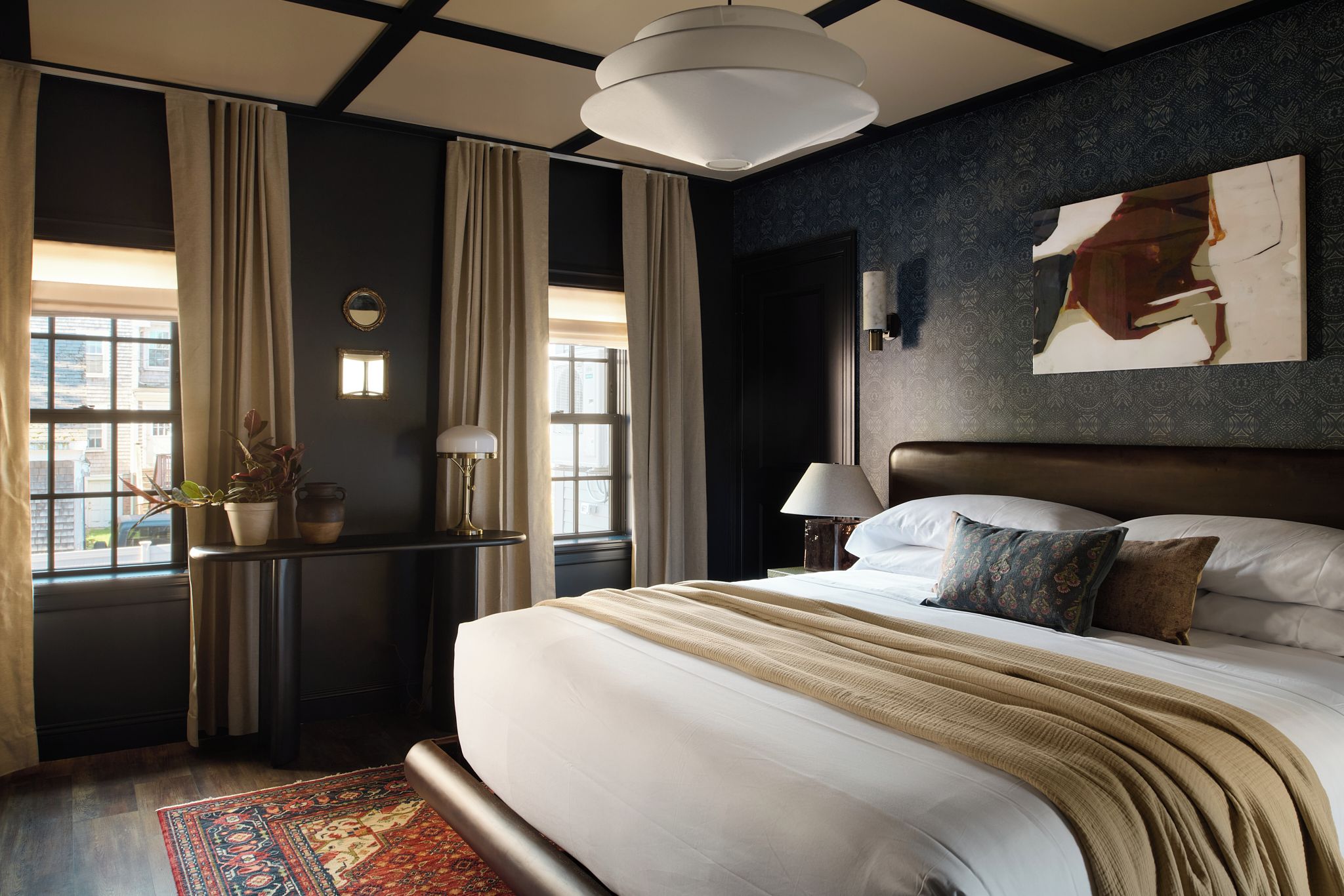 Une chambre d’hôtel avec un grand lit, un bureau avec une lampe, une fenêtre avec des rideaux et un mur orné d’un élément décoratif.
