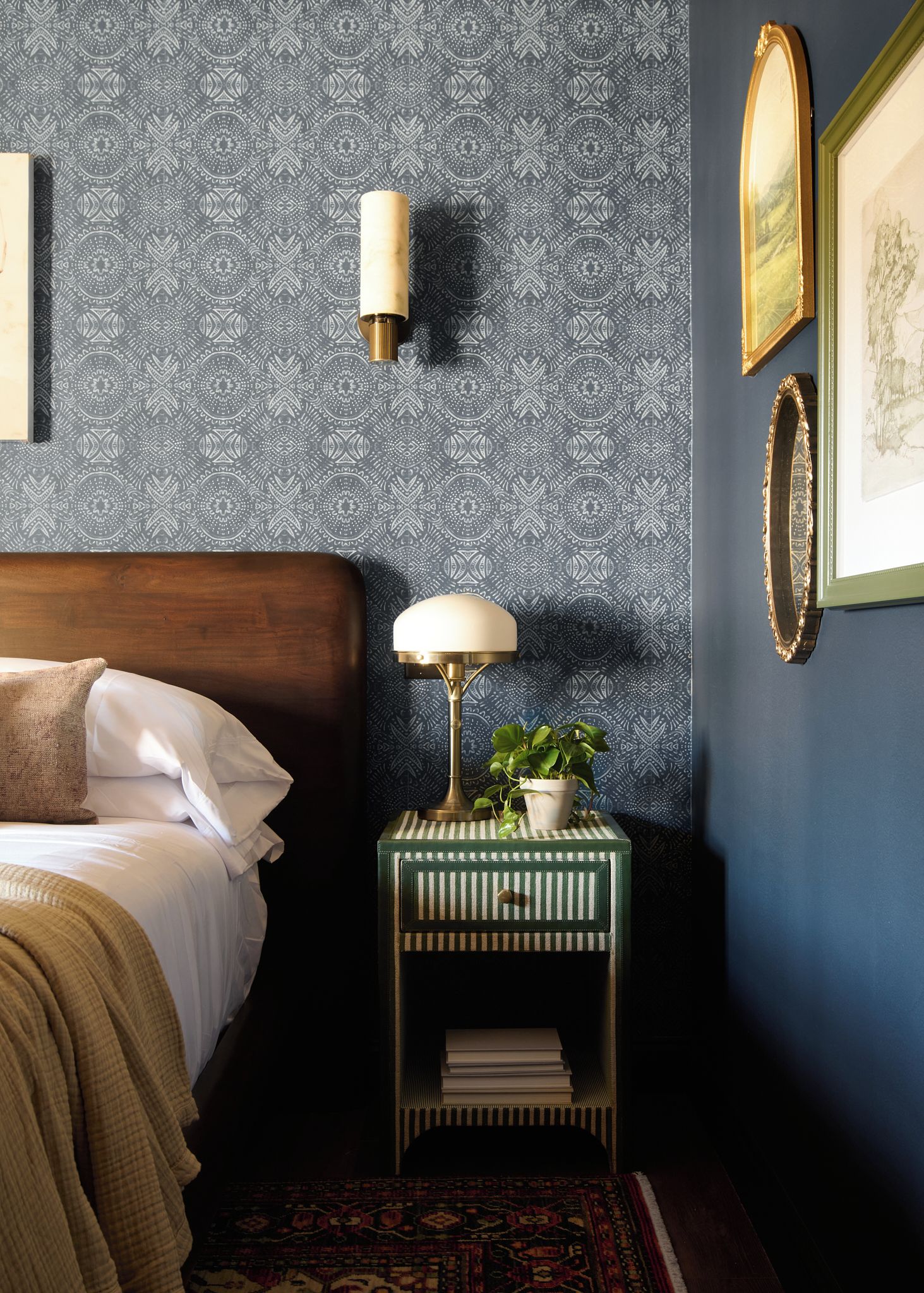 A bedroom with a patterned wallpaper, a bedside lamp, a framed picture, and a small side table with a plant.