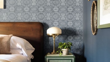 A bedroom with a patterned wallpaper, a bedside lamp, a framed picture, and a small side table with a plant.
