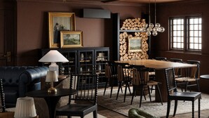 A room with a dark brown wall, a black leather sofa, a wooden dining table with chairs, a lamp, and framed pictures. - The Drew Hotel (Plymouth)