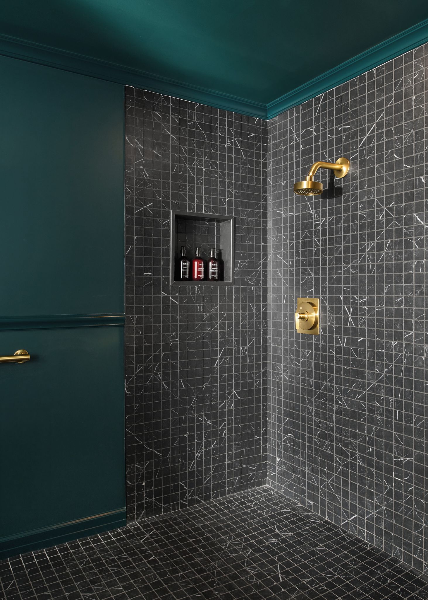 Un baño con pared de azulejos oscuros, un cabezal de ducha dorado y un estante con botellas.