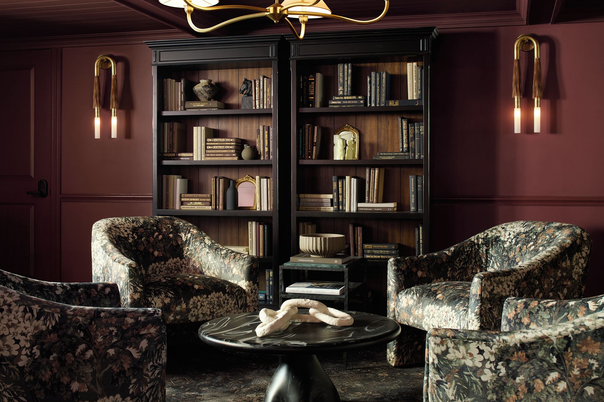 Ein Zimmer mit einem dunklen Holzregal voller Bücher, zwei sesseln mit floralem Muster und einem runden Couchtisch mit einem dekorativen Gegenstand.