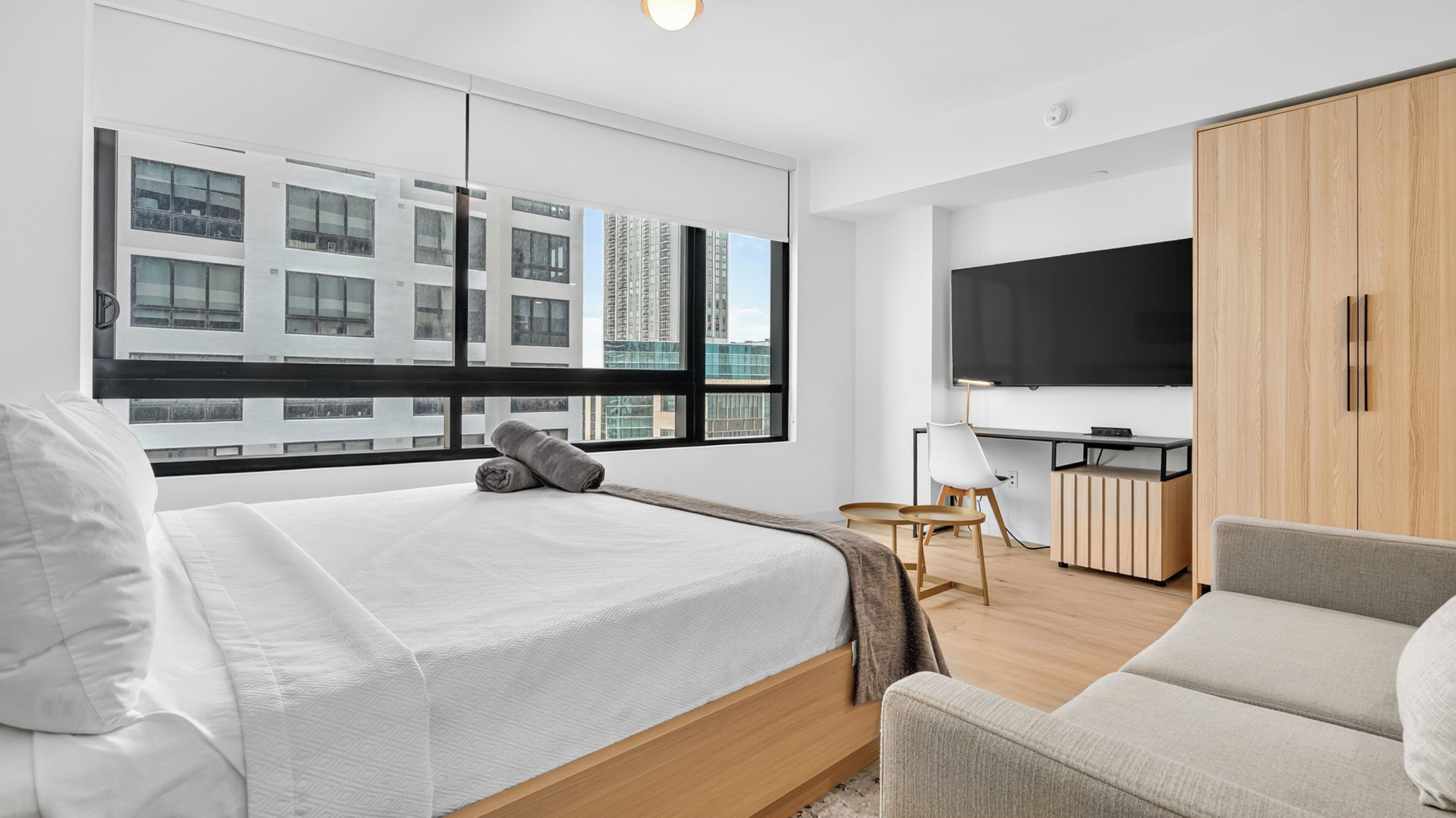 A modern hotel room with a bed, a desk, a TV, and a view of buildings.