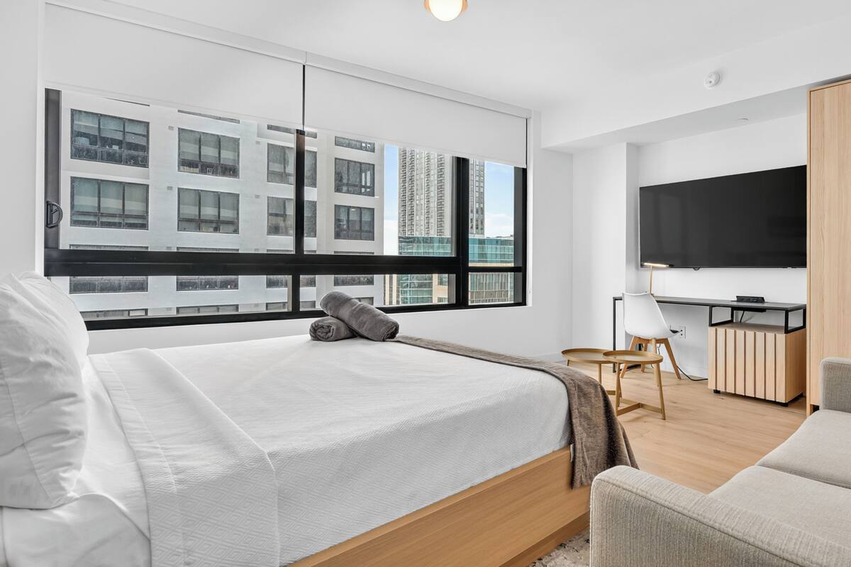 A modern hotel room with a bed, a desk, a TV, and a view of buildings.