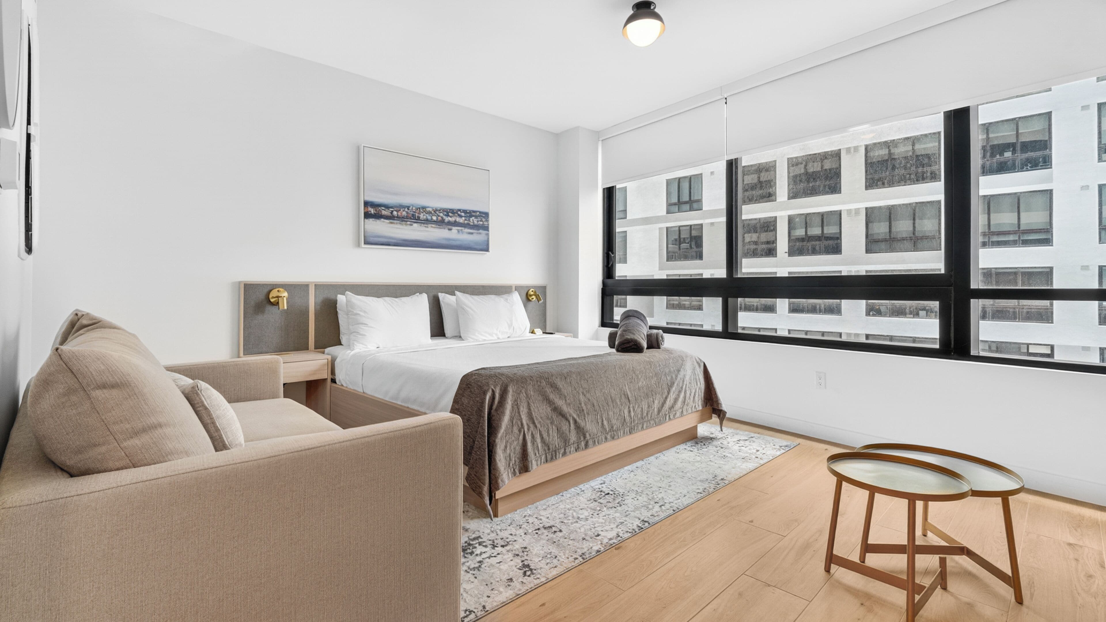 A modern hotel room with a bed, a sofa, a small table, and a large window with a city view.