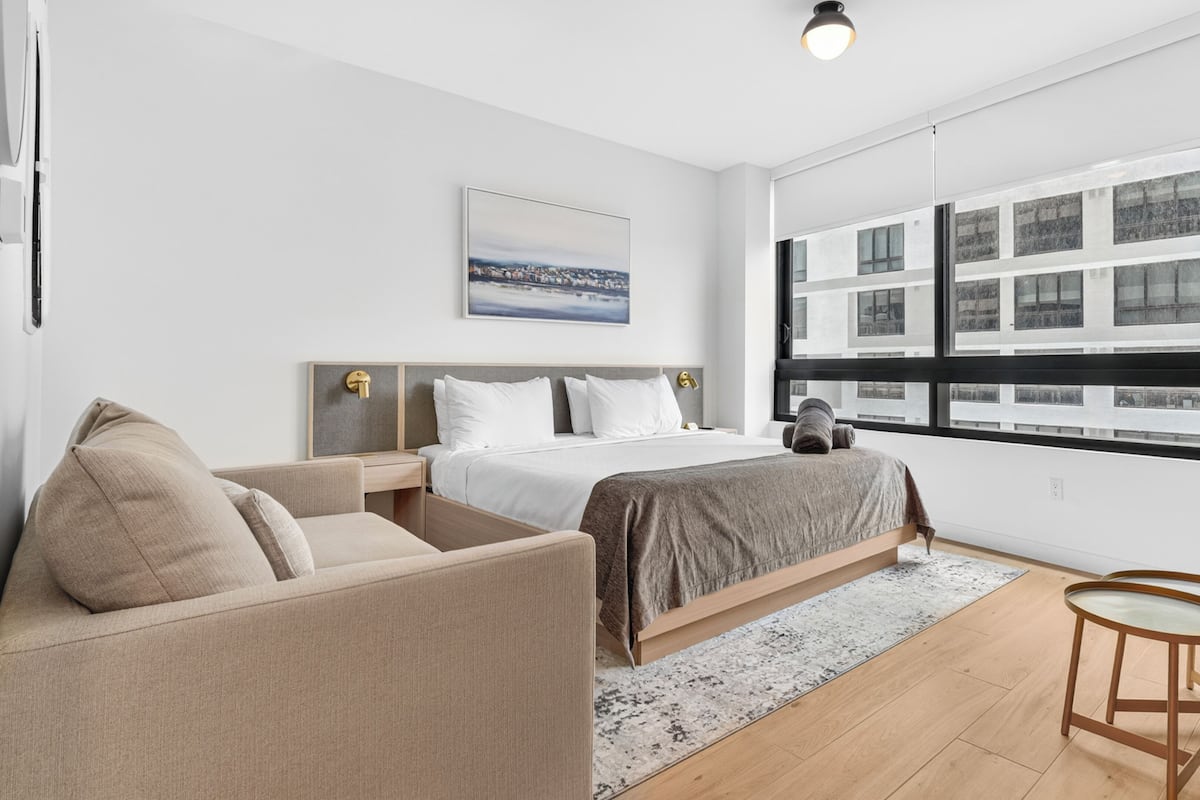 A modern hotel room with a bed, a sofa, a small table, and a large window with a city view.