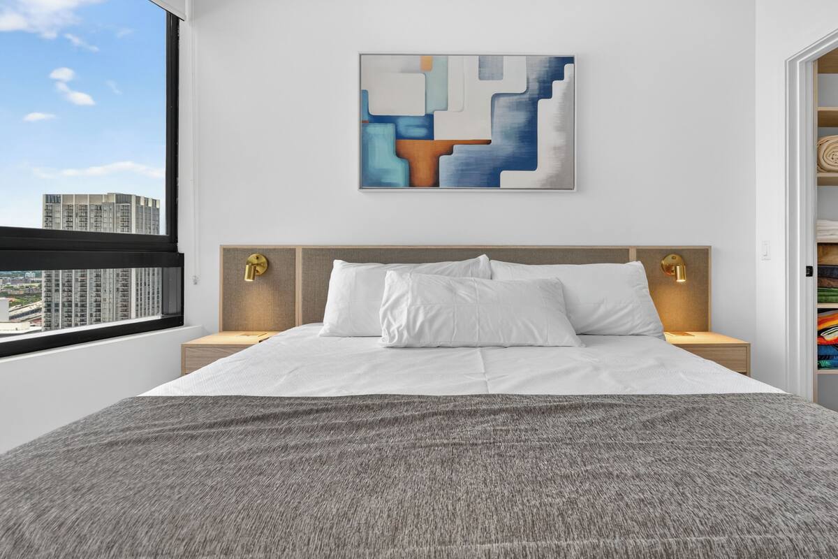 A modern hotel room with a large bed, a city view, a wall art piece, and a neatly organized wardrobe.