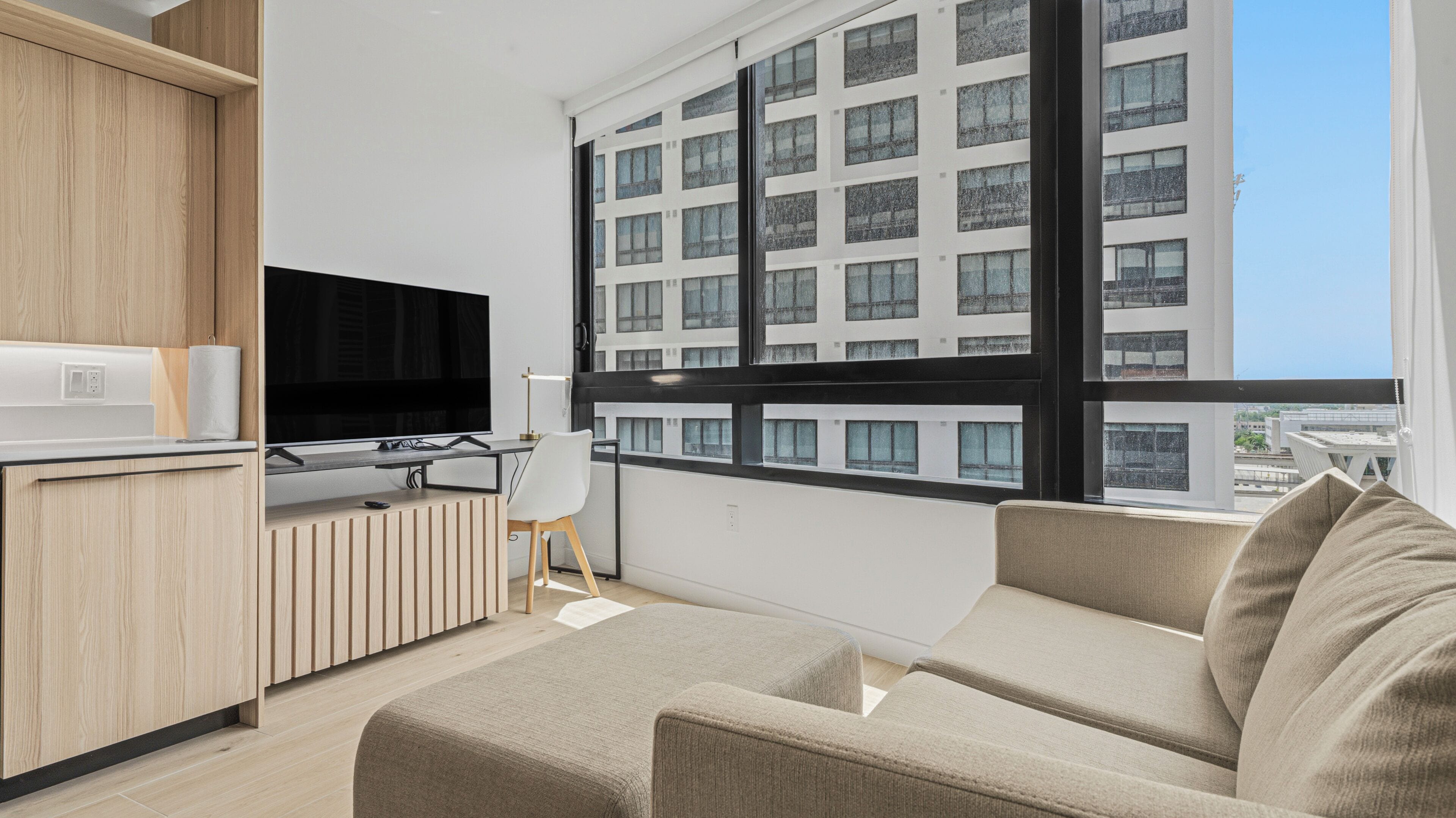 Ein modernes Wohnzimmer mit Fernseher, einer Couch, einem Schreibtisch mit Stuhl und einem großen Fenster mit Blick auf ein Gebäude.