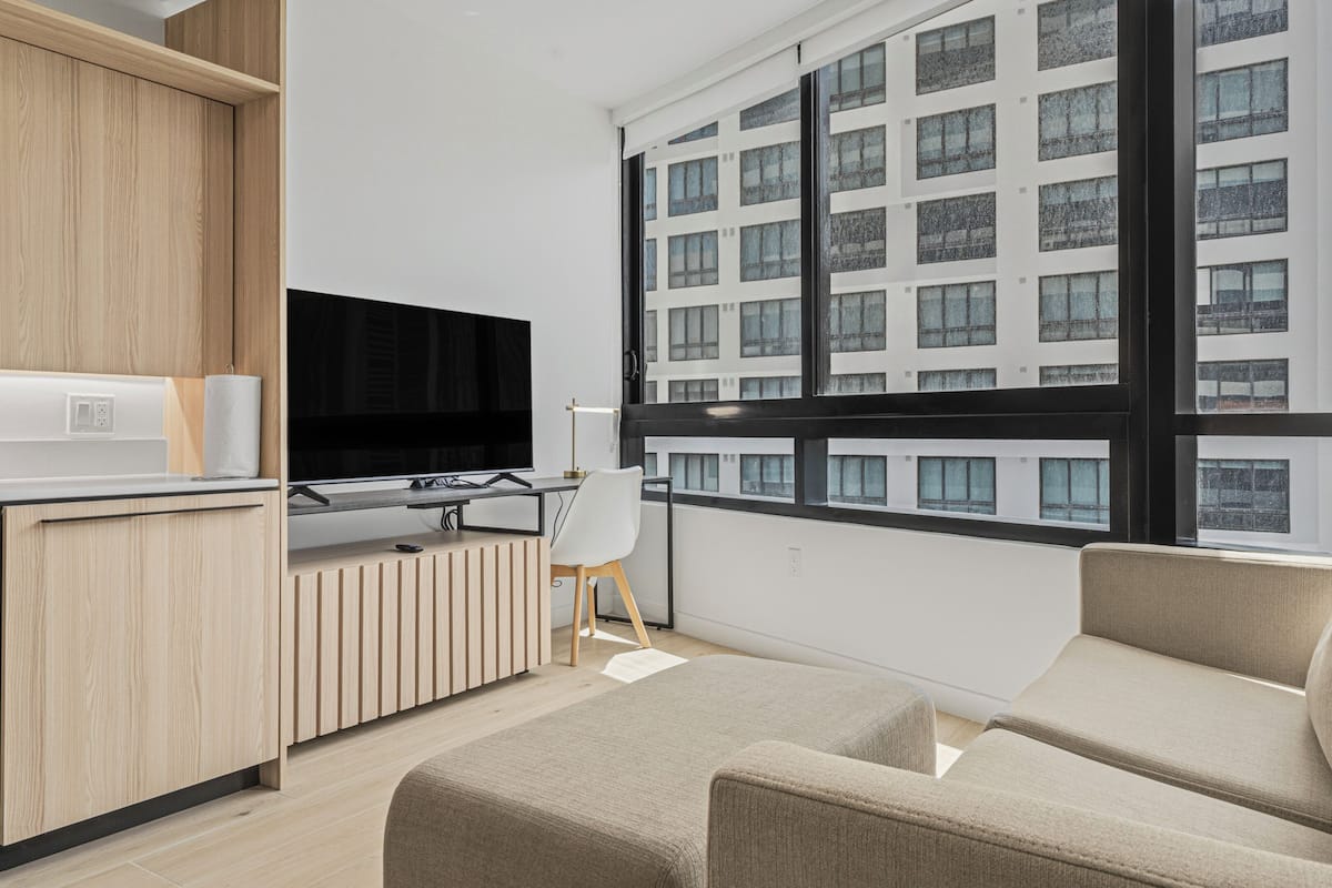 Ein modernes Wohnzimmer mit Fernseher, einer Couch, einem Schreibtisch mit Stuhl und einem großen Fenster mit Blick auf ein Gebäude.