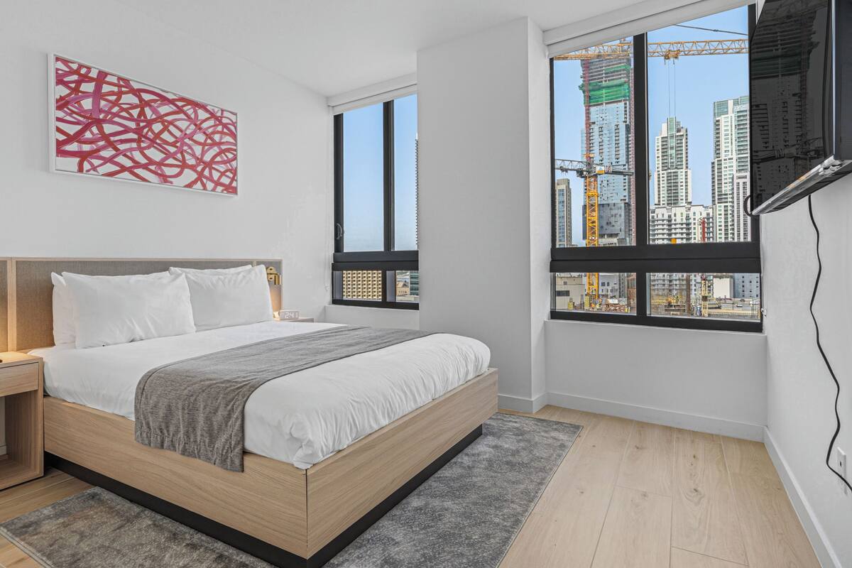 A modern bedroom with a bed, bedside tables, a TV, and large windows with a city view.