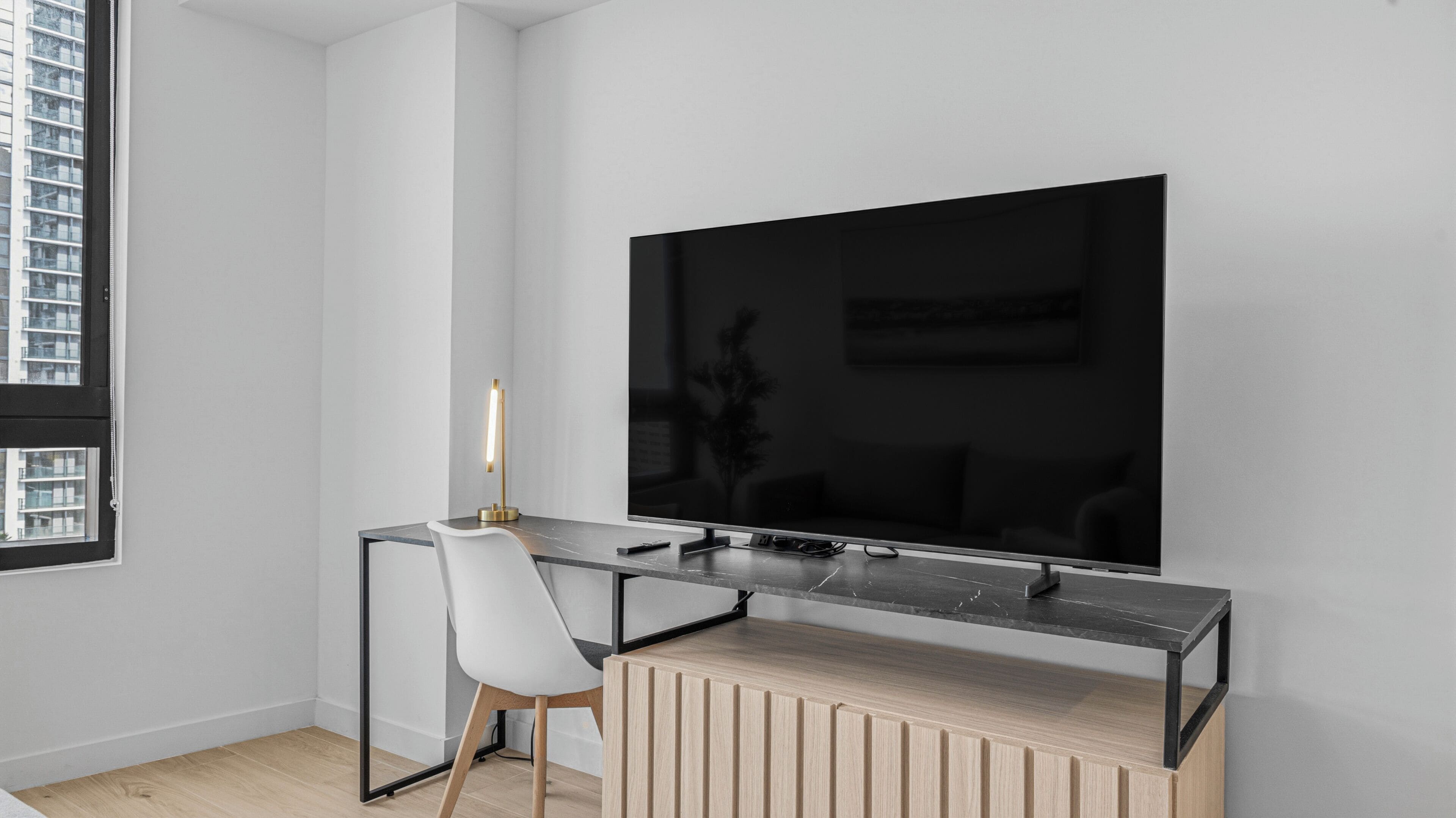 Een moderne kamer met een grote flatscreen televisie aan de muur, een bureau met glazen blad en een stoel, en een houten kast eronder.