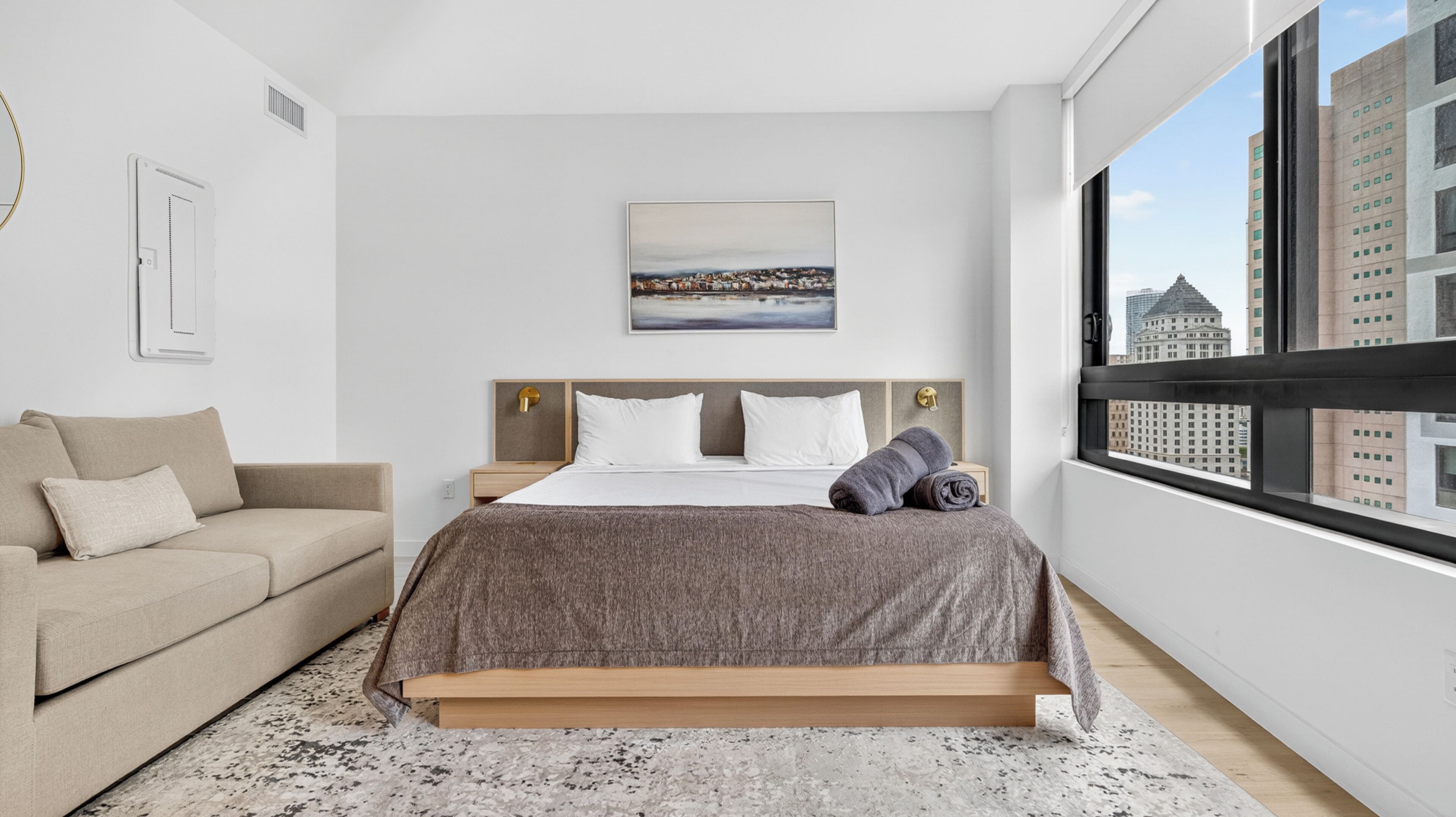 A modern bedroom with a bed, a sofa, a painting, and a city view.
