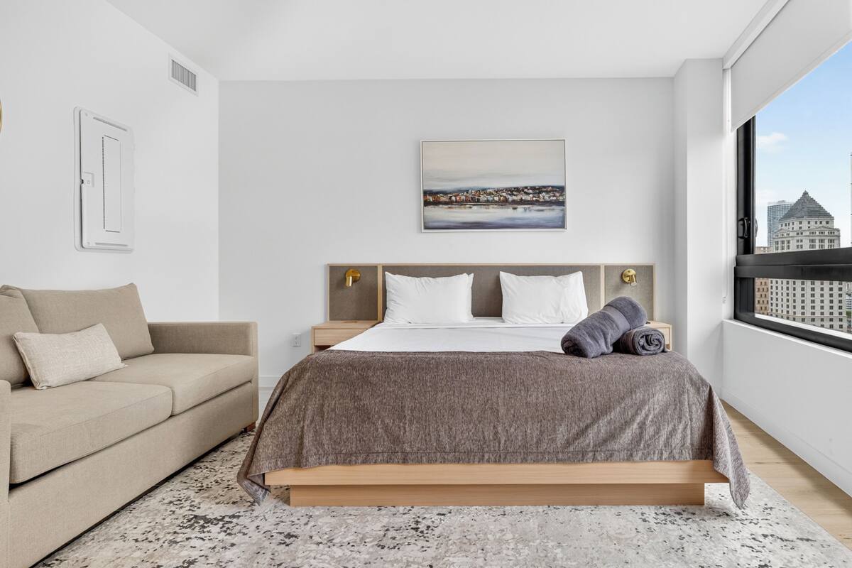 A modern bedroom with a bed, a sofa, a painting, and a city view.