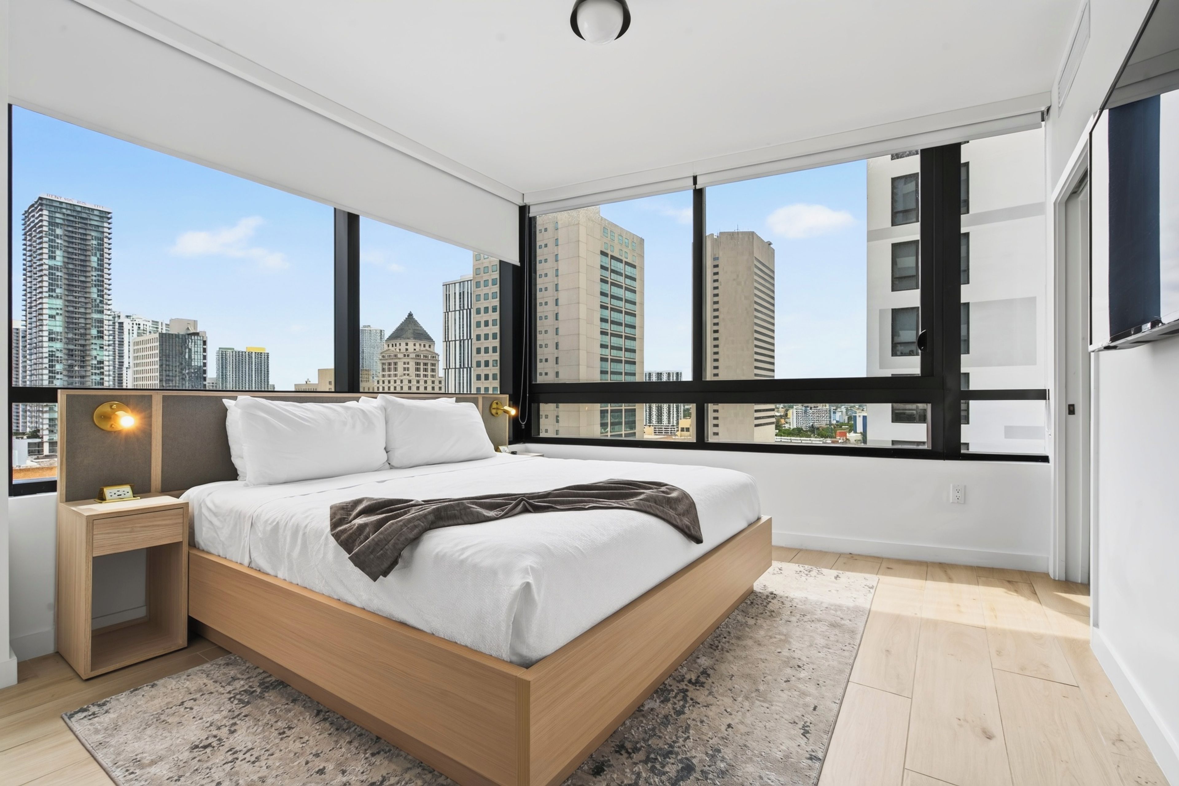 A modern bedroom with a large bed, a nightstand, a wall-mounted lamp, and a view of the cityscape through large windows.