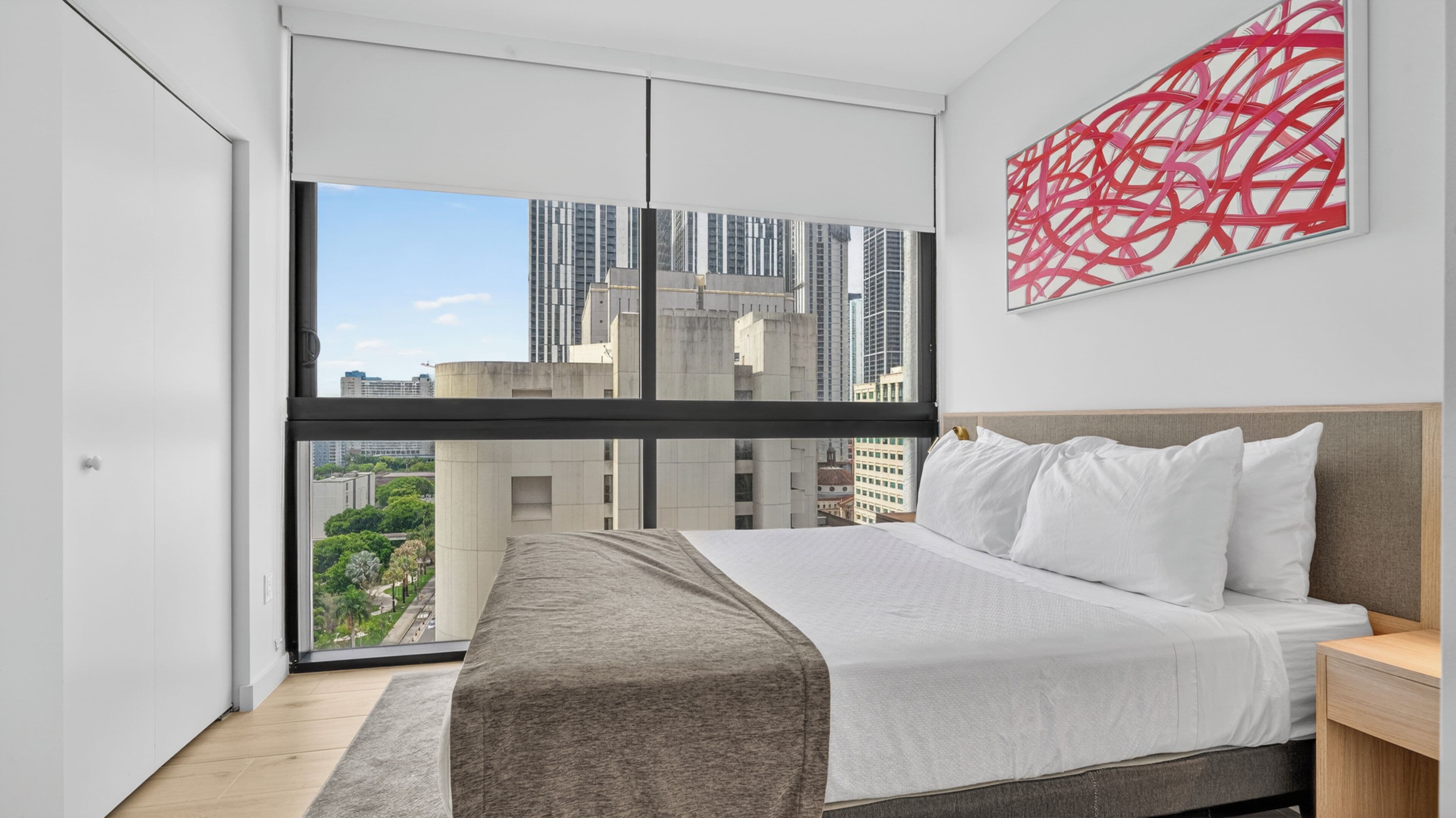 Una camera d'hotel moderna con un letto grande, un comodino e vista sulla città attraverso ampie finestre a tutta parete.