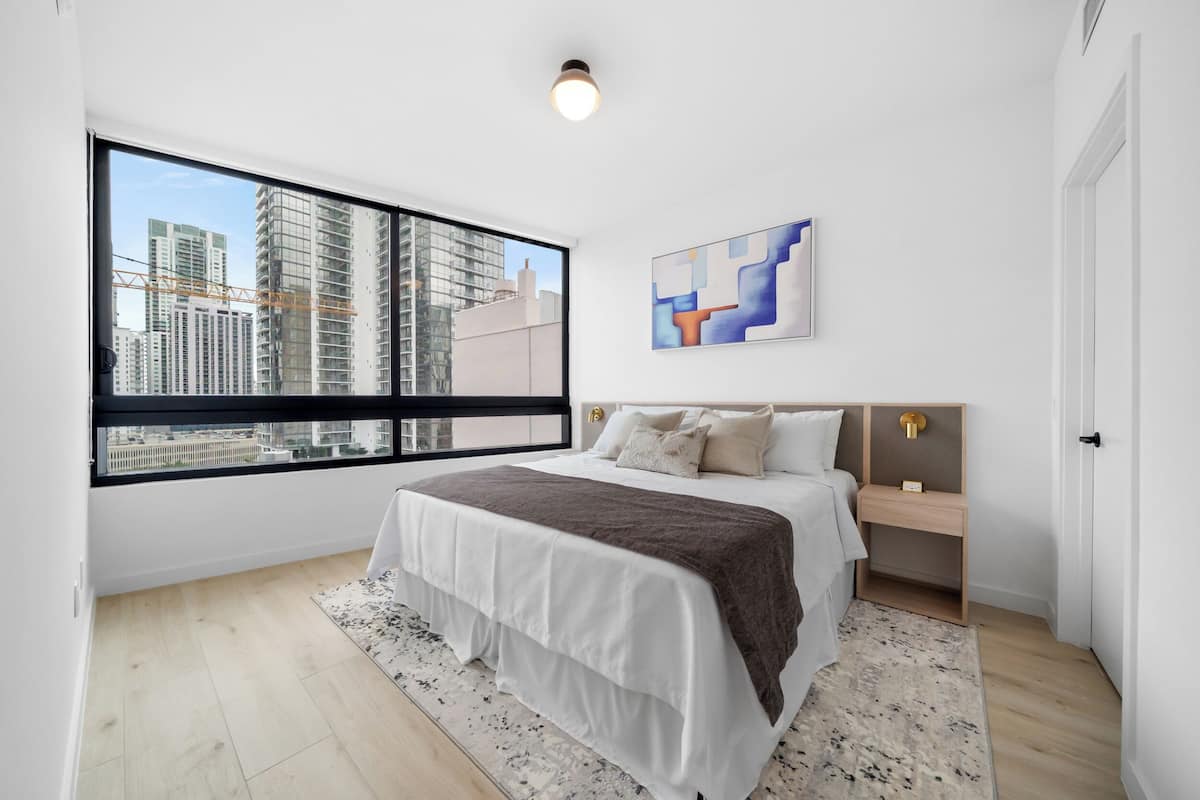 Un dormitorio moderno con un ventanal grande, una cama con cubrecama blanco y marrón, una mesita de noche de madera y un cuadro abstracto colgado en la pared.
