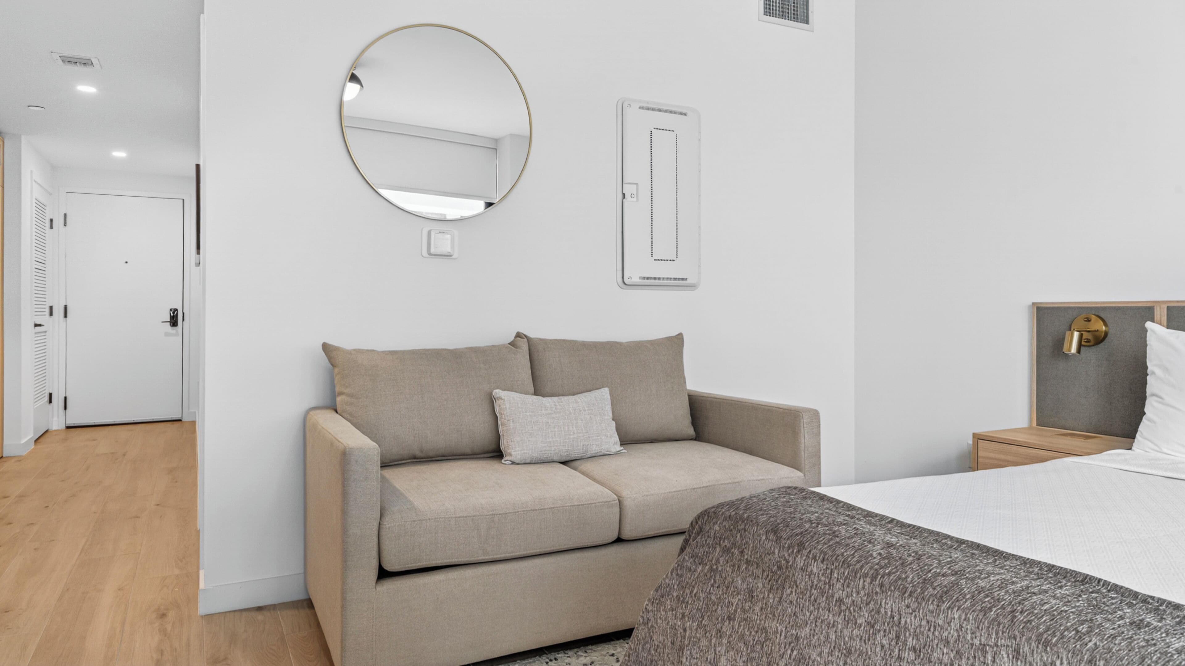 A neutral-colored room with a sofa, a bed, a round mirror, and a wall-mounted light fixture.