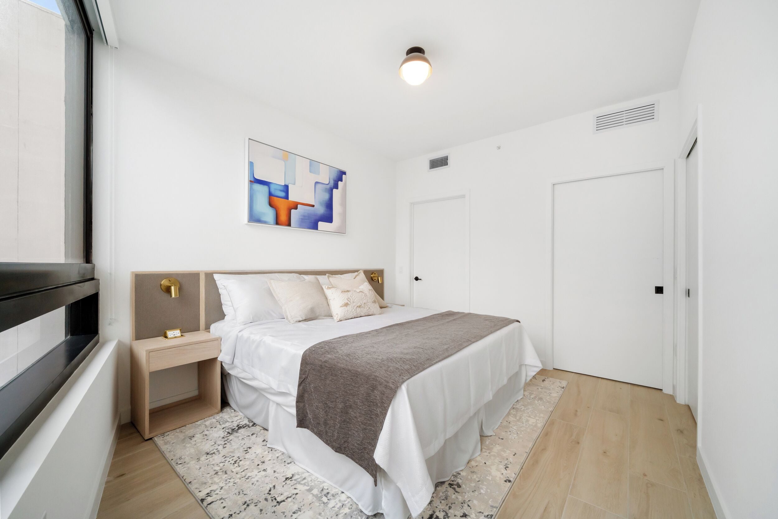 A bedroom with a bed, bedside tables, a nightstand, a painting, a door, and a window.