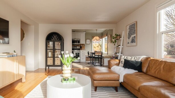 A modern living room with a brown leather sofa, a white coffee table, and a dining area in the background. - The Ballard Vista | Sleeps 6 + Patio & Fire Pit (Seattle)