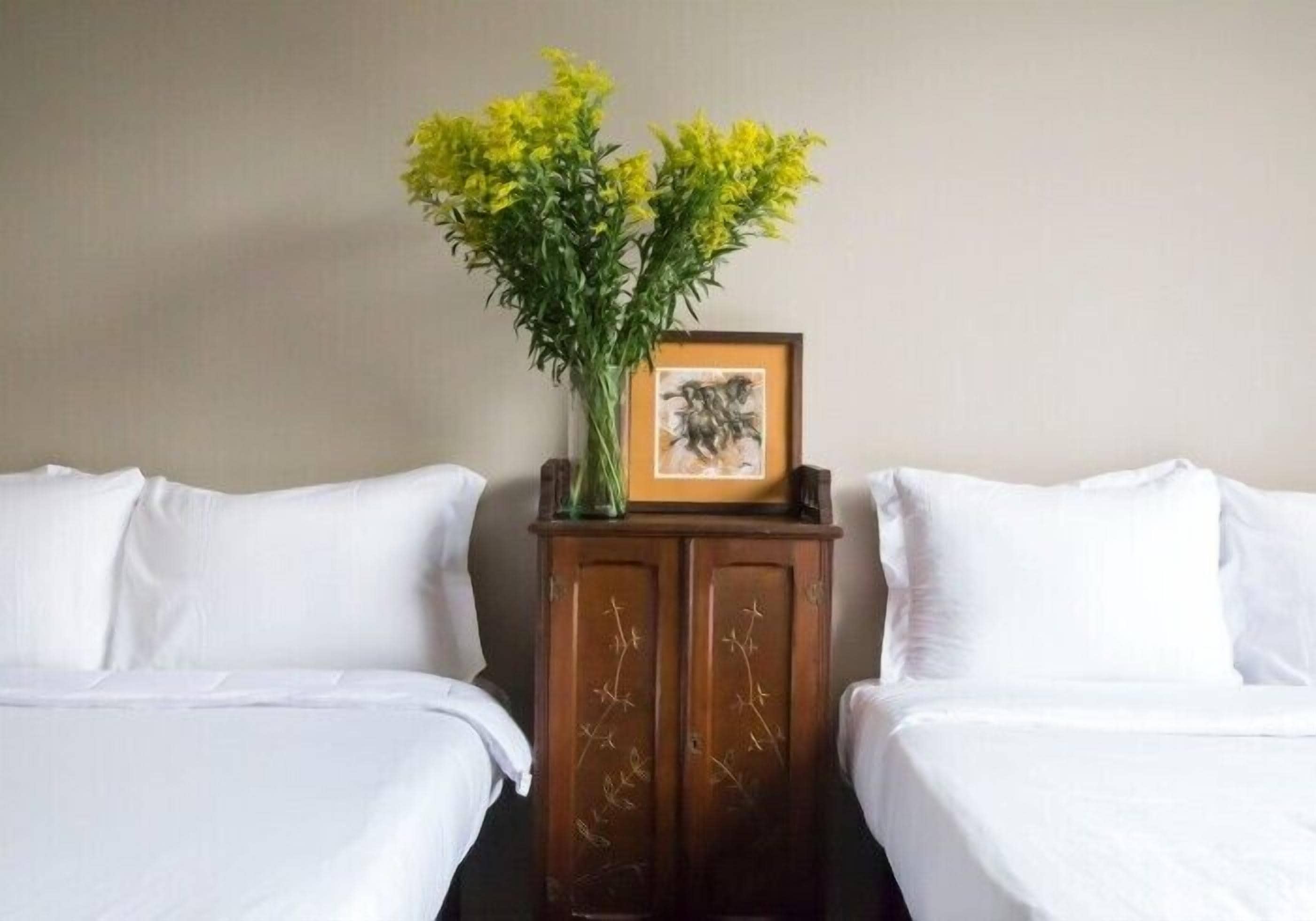 Une chambre d’hôtel avec deux lits, une table de chevet en bois ornée d’un vase de fleurs jaunes et un tableau encadré.