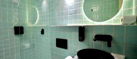 Egy fürdőszoba, melyben üvegfalas zuhanyzó, kerek tükör, fekete mosdókagyló és WC található.