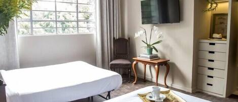 Ein Hotelzimmer mit zwei Betten, einem Fernseher, einem kleinen Tisch mit Frühstücksbrettern, einem Sessel und einem Fenster mit Vorhängen.