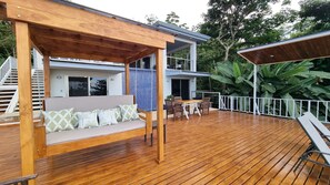 Una terraza de madera con una silla mecedora y una mesa de comedor, rodeada por una valla blanca y abundante vegetación.