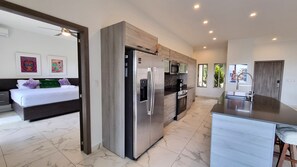 Una cocina moderna con refrigerador empotrado, microondas y cafetera. Hay una zona de comedor con una isla grande y taburetes de bar. La habitación tiene una cama grande con cabecera y dos cuadros colgados.