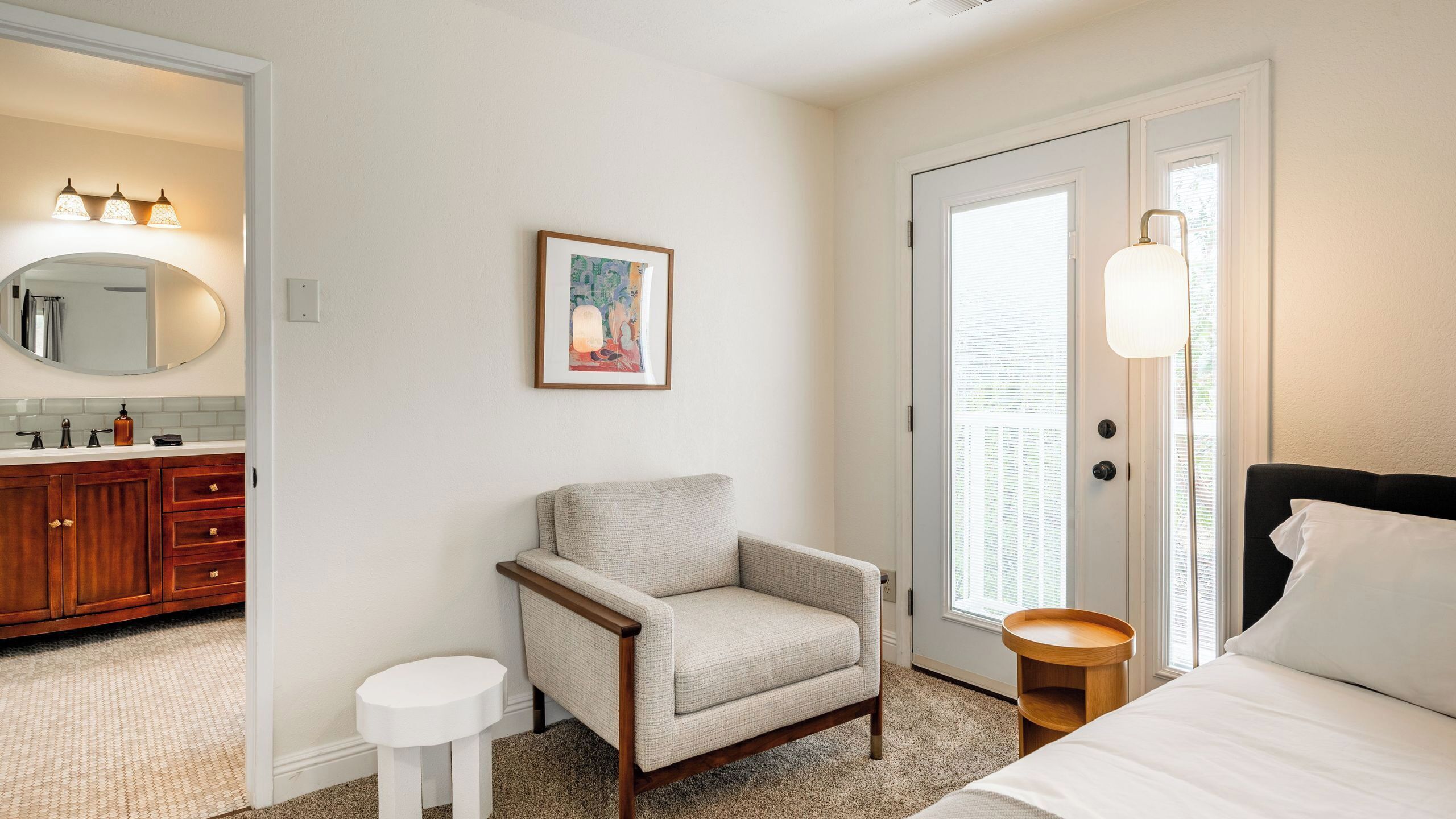 A bedroom with a bed, a chair, a round wooden side table, a framed picture on the wall, and a bathroom with a mirror, sink, and towel rack.
