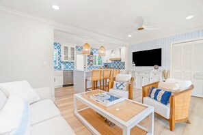 Ein modernes Wohnzimmer mit einem weißen Sofa, einem Couchtisch und Korbstühlen. Im Hintergrund ist eine Küche mit blauen und weißen Fliesen und einem Flachbildfernseher zu sehen.