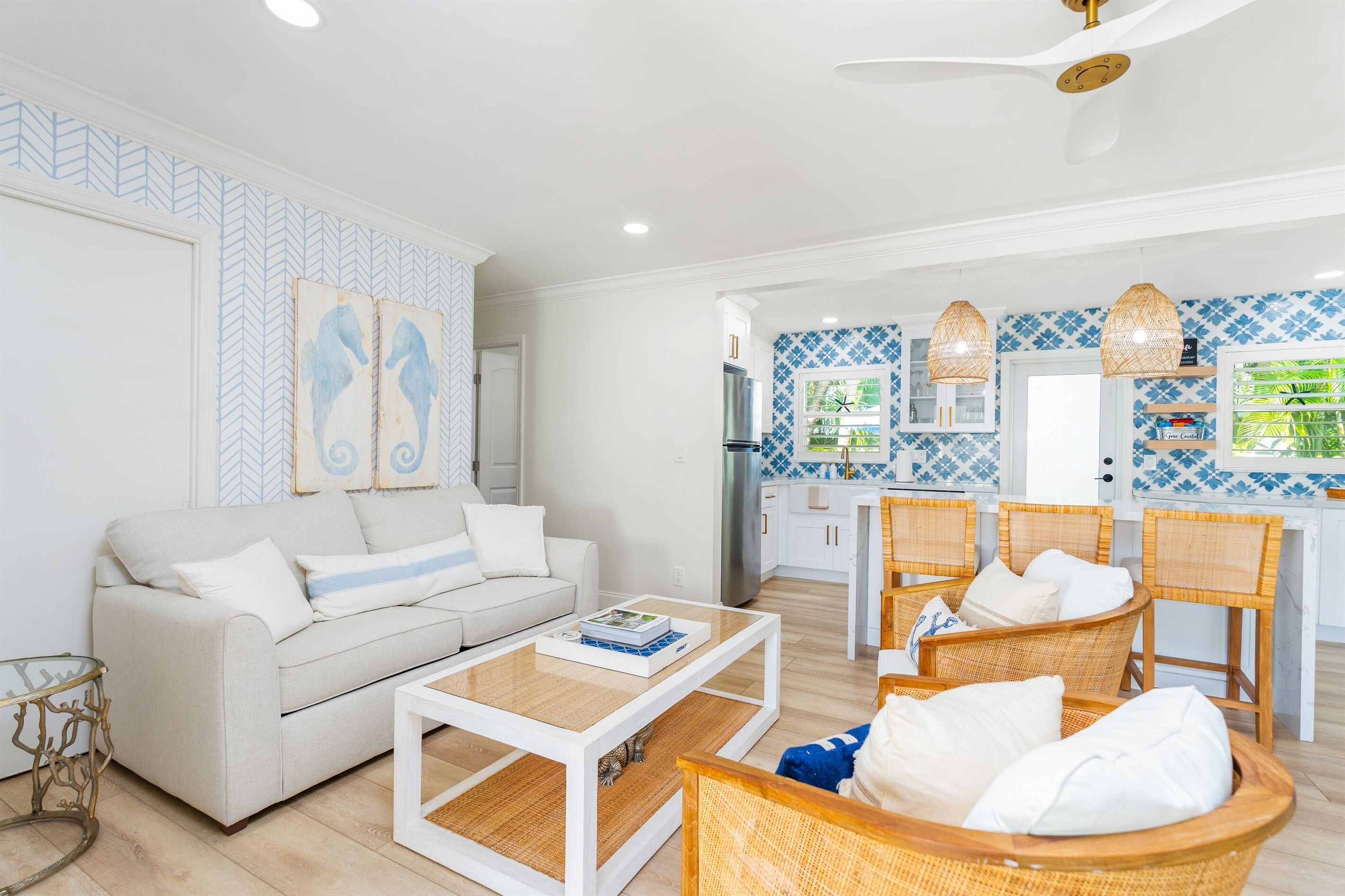 A modern living room with a sofa, coffee table, and wicker chairs. A kitchen with blue and white tiles and wicker pendant lights is visible in the background.