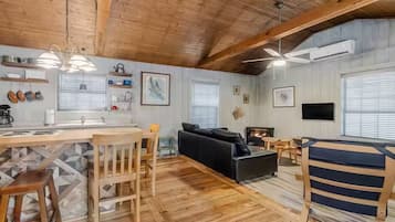Interior acogedor de una cabaña con techo de madera, una zona de cocina con barra, un sofá negro y un televisor.