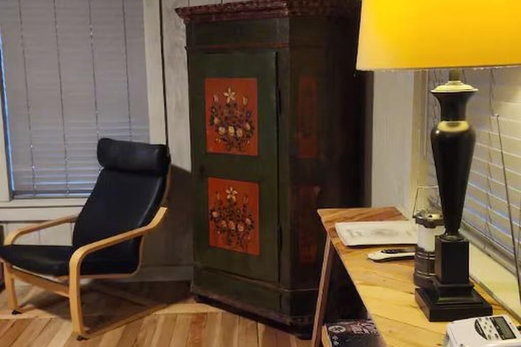 Een kamer met een houten vloer, een zwarte stoel, een groen kastje met decoratieve panelen en een tafel met een lamp en een afstandsbediening.