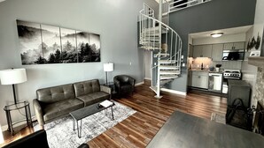Ein modernes Wohnzimmer mit einem grauen Sofa, einem Couchtisch, einem schwarzen Sessel und einem Gemälde an der Wand. Eine Wendeltreppe führt zu einer Küchenzeile mit Mikrowelle und Herd.