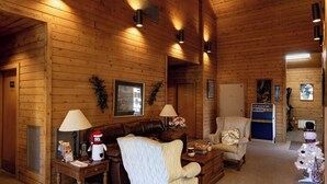 Ein gemütliches Holzblockhaus-Interieur mit Holzwänden, Kamin, einer Couch, einem Sessel und einem kleinen Tisch mit Lampe.