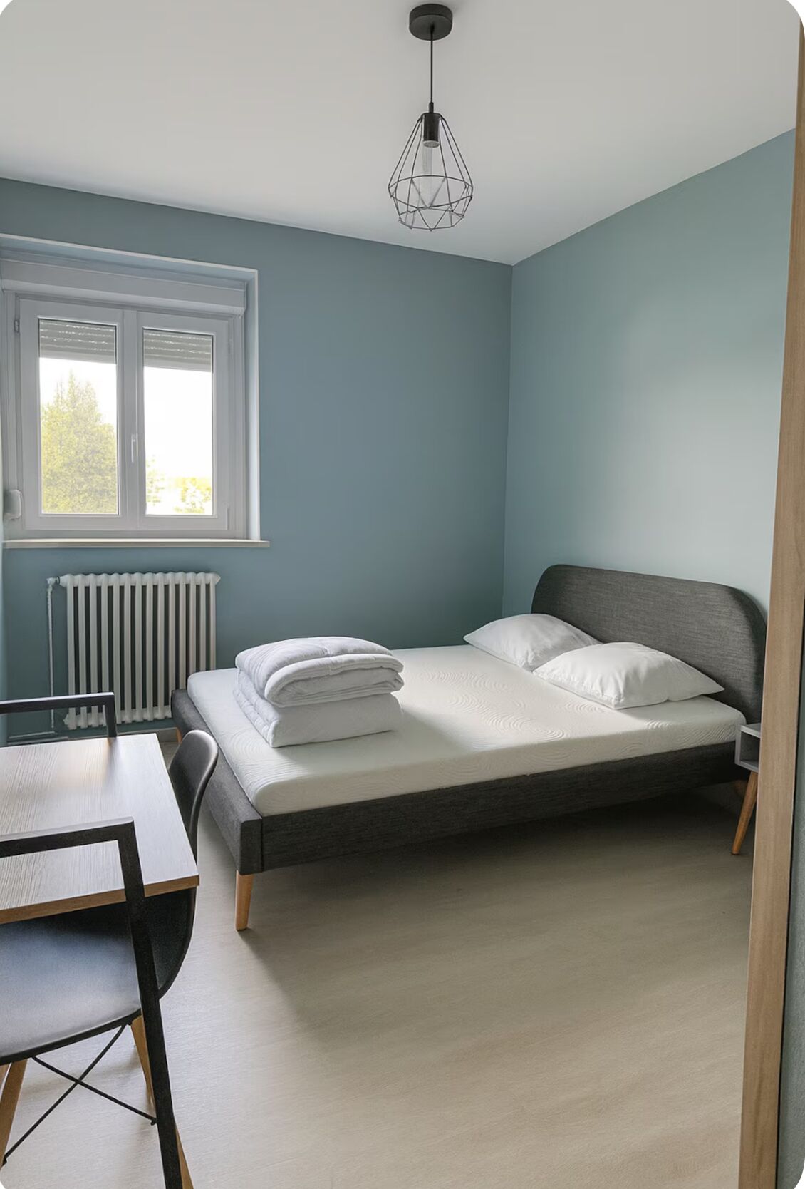 Une chambre à coucher avec un lit, une table et une chaise. Il y a une fenêtre avec des stores et un radiateur en dessous.