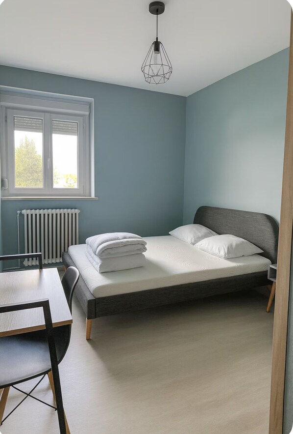 Un dormitorio con cama, una mesa y una silla. Se ve una ventana con persianas y un radiador debajo.