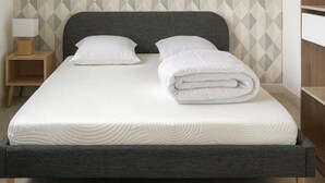 A neatly made bed with a grey headboard and white bedding, a wooden nightstand with a lamp, and a geometric patterned wall.