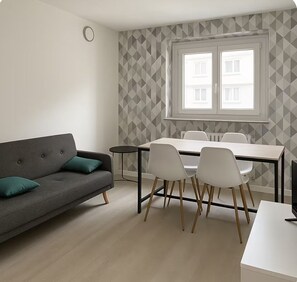 Una sala moderna con un sofá gris, una mesa de comedor con cuatro sillas y un ventanal con vistas a edificios.