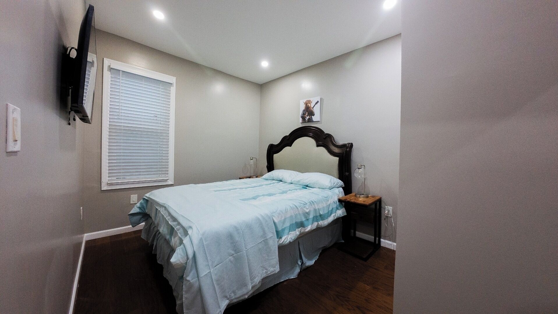 A bedroom with a bed, a nightstand, a wall-mounted TV, and a window with blinds.
