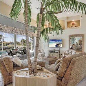 A living room with a large sofa, a coffee table with a palm tree centerpiece, a flat-screen TV, and a dining area with outdoor seating. - 400 Bald Eagle Drive, centrally located spacious home with a gorgeous pool area! (Marco Island)