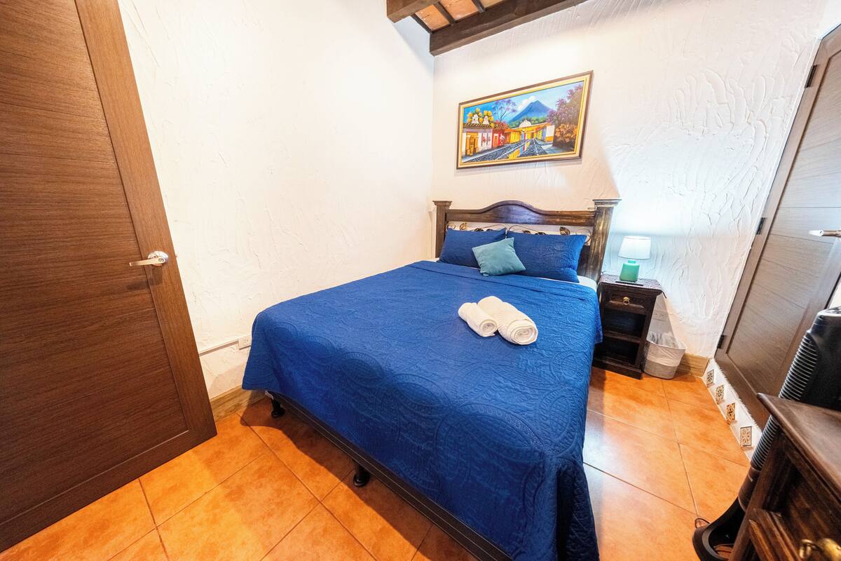 Un dormitorio con una cama, una mesita de noche, una lámpara, un cuadro en la pared y una puerta de madera.
