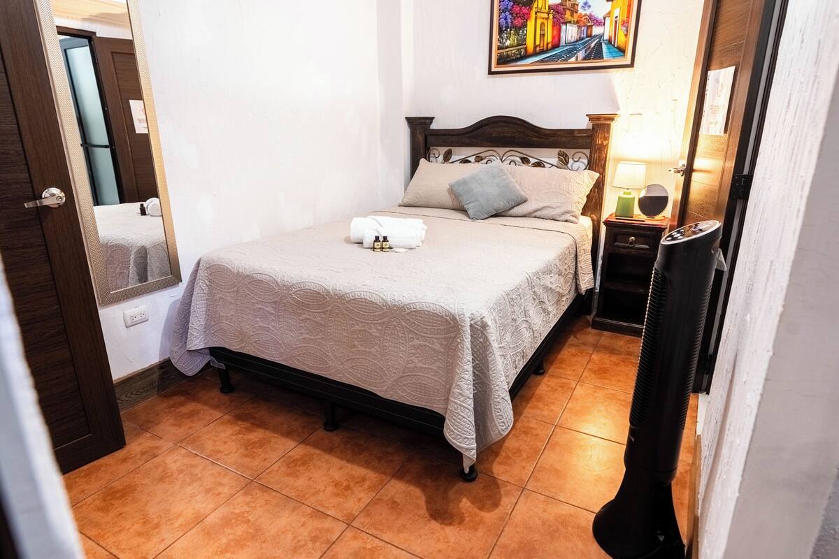 Una cama bien tendida con una colcha blanca, una mesita de noche con una lámpara, y un cuadro colgado en la pared.