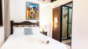 Quarto de hotel com uma cama, uma mesa de cabeceira com abajur, um quadro na parede e uma porta que dá para o banheiro.