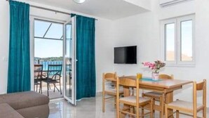 Quarto com mesa de jantar de madeira, cadeiras, sofá e televisão. Há uma porta de vidro deslizante que dá para uma área externa.