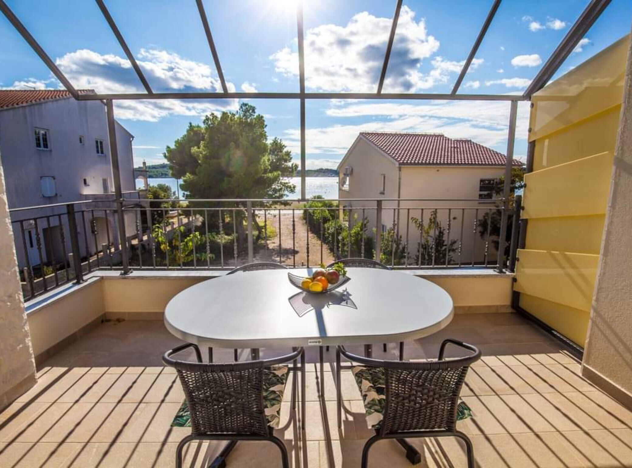 Een balkon met een ronde tafel en twee stoelen, met uitzicht op een kustgebied met gebouwen en bomen.